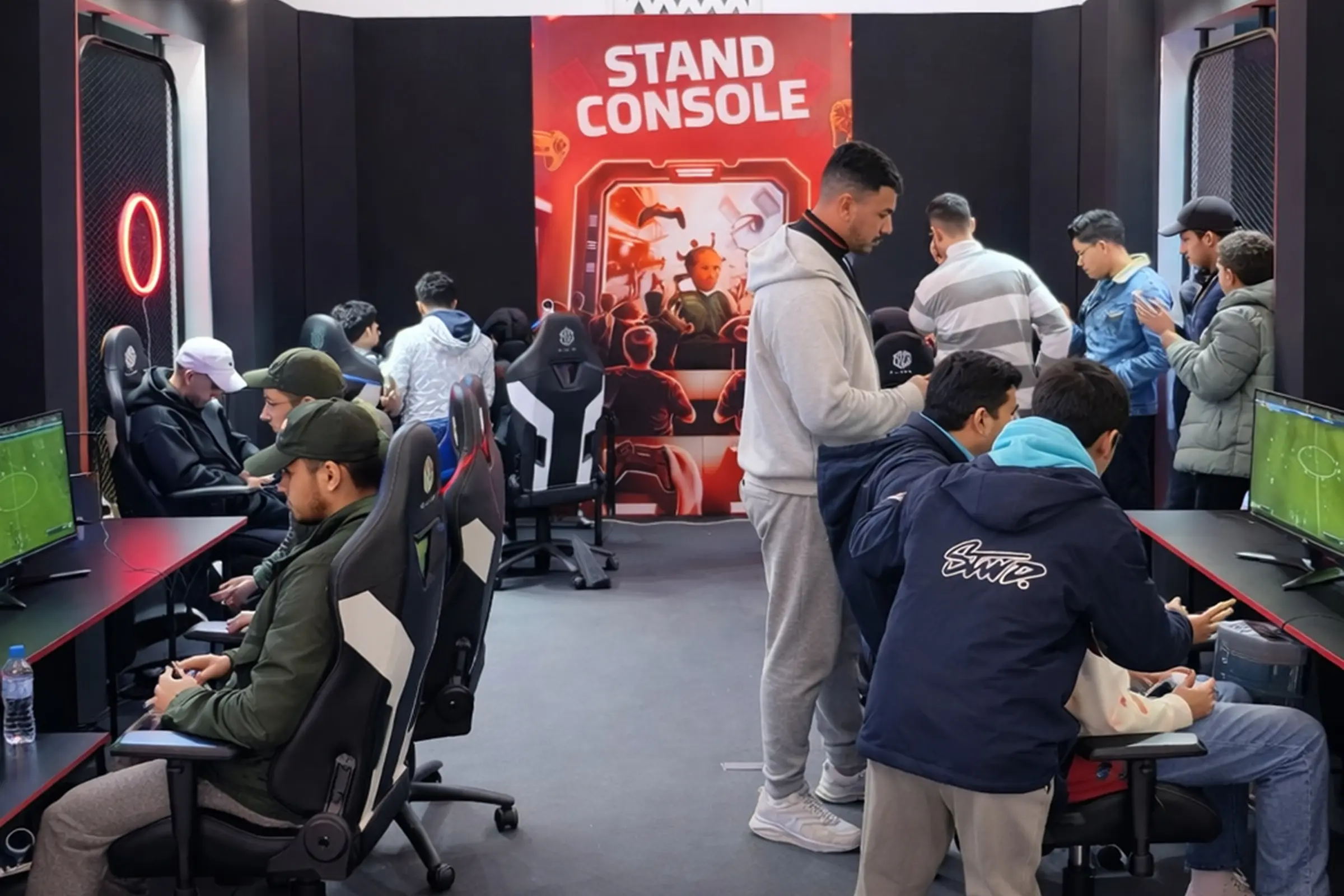 Stand console sur mesure de la Fédération Royale Marocaine des Jeux Électroniques (FRMJE) dans l’eZone Fanzone à Casablanca lors de la CAN 2025, avec des joueurs e-sport en compétition sur consoles et PC, attirant un large public dans un espace gaming dédié.